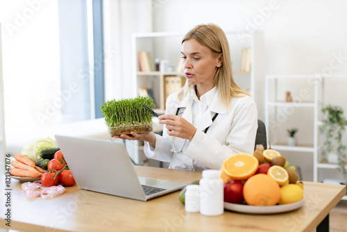 Smiling Caucasian woman introducing microgreens during online conversation with client via laptop webcam. Modern nutrition professional advising young vegetable sprouts for healthy eating.