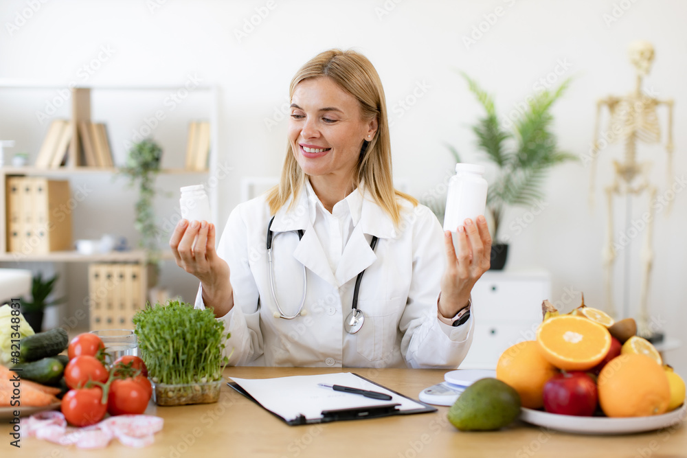 Portrait of positive Caucasian lady in lab coat smiling at camera while demonstrating two pills bottles in office. Cheerful expert in food proposing dietary supplements to improve healthy meals. Portrait of positive Caucasian lady in lab coat smiling at camera while demonstrating two pills bottles in office. Cheerful expert in food proposing dietary supplements to improve healthy meals.
