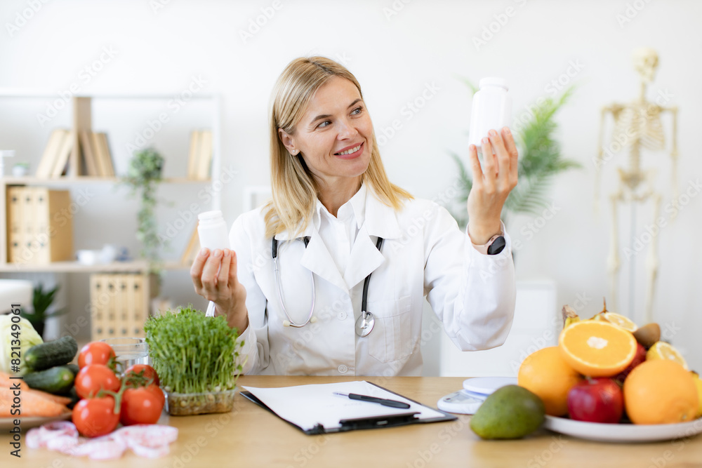 Cheerful expert in food proposing dietary supplements to improve healthy meals. Portrait of positive Caucasian lady in lab coat smiling at camera while demonstrating two pills bottles in office.