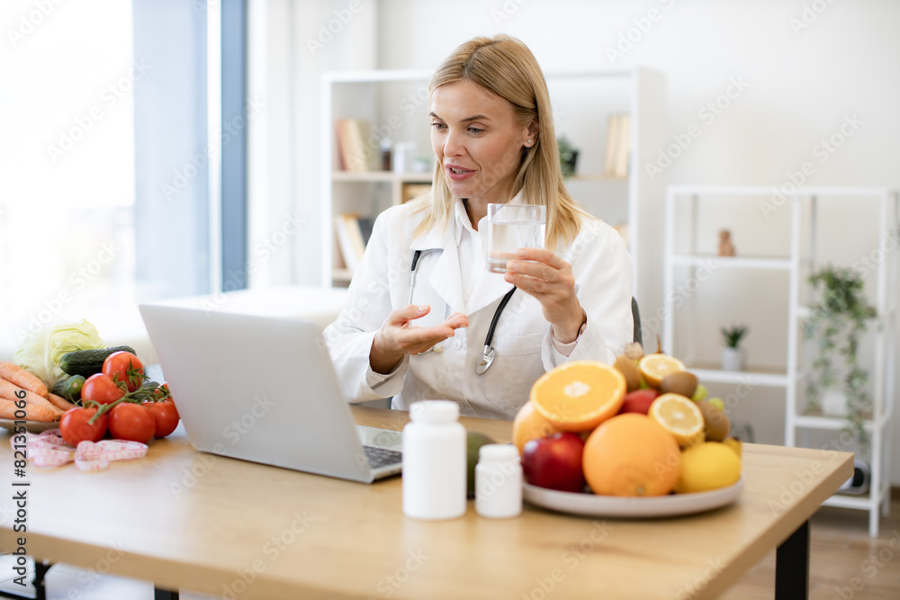 Experienced female nutritionist tells patient about benefits of water during an online session using laptop. Female doctor in white coat holding glass of water during a video call. Experienced female nutritionist tells patient about benefits of water during an online session using laptop. Female doctor in white coat holding glass of water during a video call.
