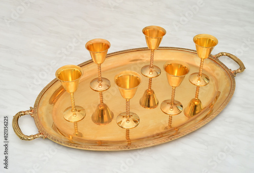 Five golden antique goblets with high stems on the golden tray on the light gray marble table texture. Selective focus.