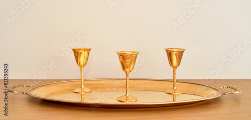 Three golden antique goblets with high stems on the golden tray on the brown wooden table. Close-up. Copy space. Selective focus.

