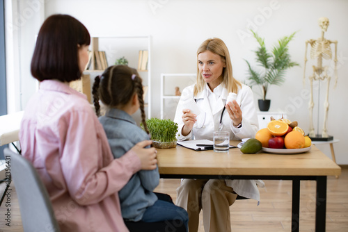 Happy pretty female nutritionist holding bottles with pills as nutrition supplements for little girl sitting on moms lap. Mature pediatrician receives female patient in modern clinic.