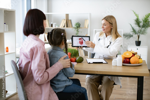 Mother with little girl at appointment with family nutritionist. Mature experienced female doctor showing tablet with gastrointestinal tract image for treatment planning to little female child. photo