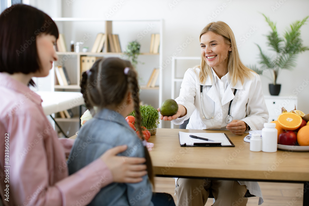 Mature woman with fresh avocado in hand chatting with little female child and her mother in consulting room. Experienced medical specialist giving advice on improving diet by abundance of nutrients. Mature woman with fresh avocado in hand chatting with little female child and her mother in consulting room. Experienced medical specialist giving advice on improving diet by abundance of nutrients.