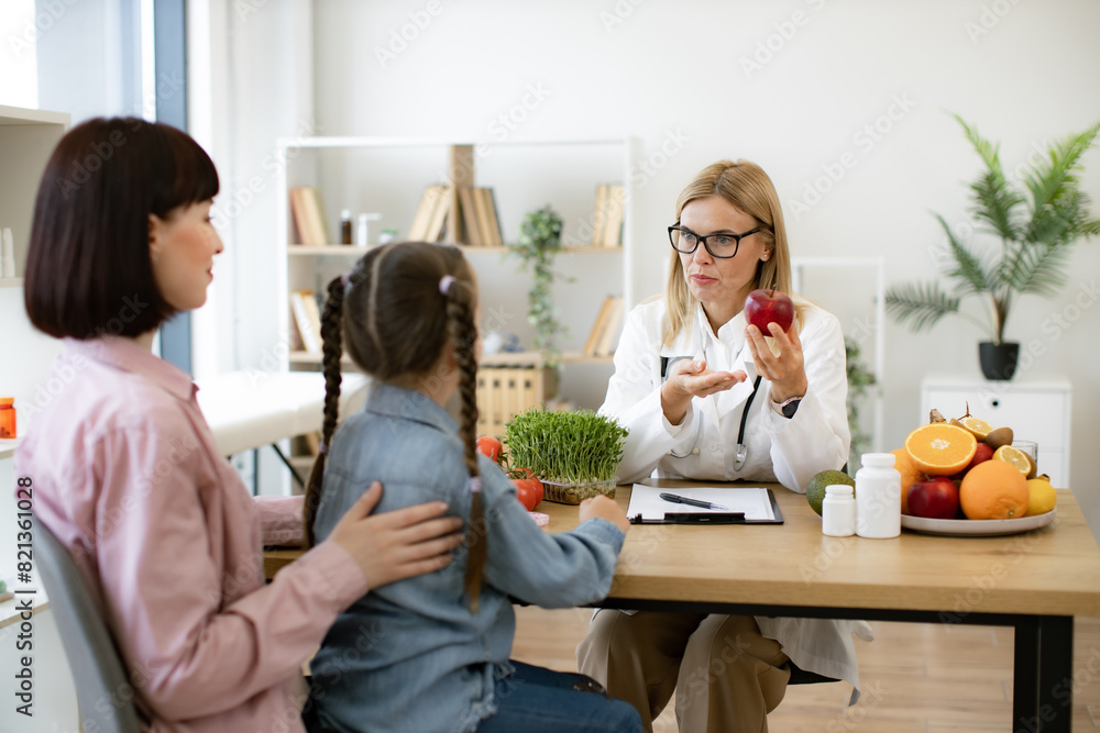 © sofiko14 - Mature woman with fresh apple in hand chatting with little female child and her mother in consulting room. Experienced medical specialist giving advice on improving diet by abundance of nutrients. © sofiko14 - Mature woman with fresh apple in hand chatting with little female child and her mother in consulting room. Experienced medical specialist giving advice on improving diet by abundance of nutrients.