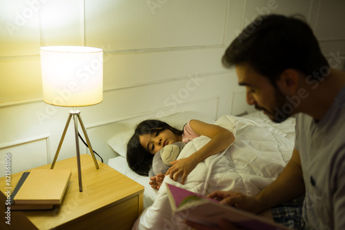 Dad reads a bedtime story to his little girl, tucked into bed at home with soft, warm lighting brightening the evening