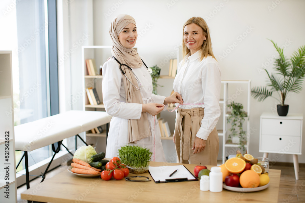 Efficient dietitian calculating body mass index for assessing healthy weight. Muslim woman in doctor's coat and hijab finding out waist length of female patient with measuring tape in consulting room. Efficient dietitian calculating body mass index for assessing healthy weight. Muslim woman in doctor's coat and hijab finding out waist length of female patient with measuring tape in consulting room.