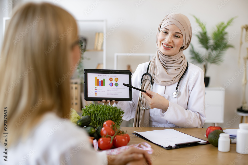 © sofiko14 - Mature lady patient visiting arabian woman nutritionist in hijab at modern clinic. Muslim female doctor showing on tablet graphs of food for healthy eating.