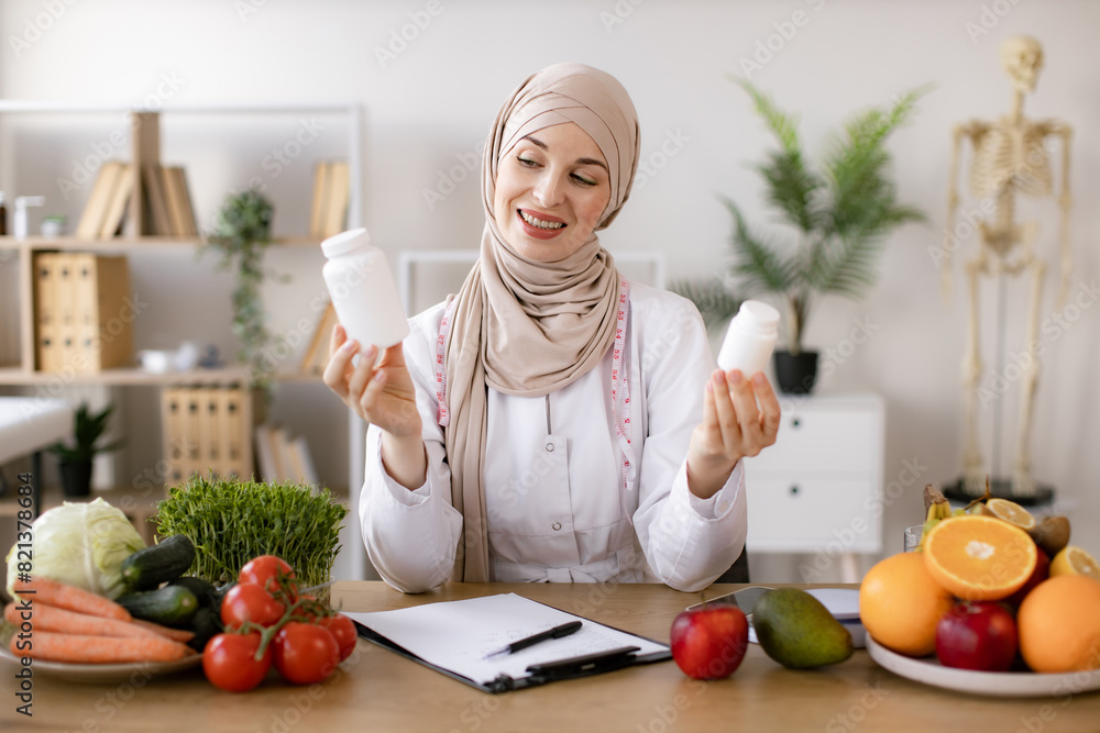 Cheerful expert in food proposing dietary supplements to improve healthy meals. Portrait of positive Muslim lady in hijab and lab coat smiling at camera while demonstrating two pills bottles in office Cheerful expert in food proposing dietary supplements to improve healthy meals. Portrait of positive Muslim lady in hijab and lab coat smiling at camera while demonstrating two pills bottles in office
