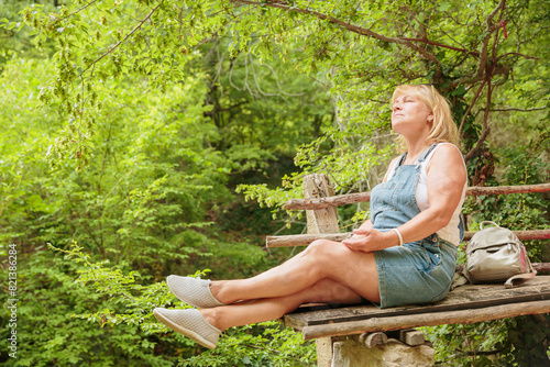 Mature woman sitting on wooden bench in summer forest. Attractive female traveler relaxing, meditation, breathing and enjoying nature on hiking trail. Concept of mental detox and physical health