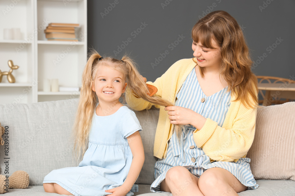 Nanny brushing little girl's hair on sofa at home