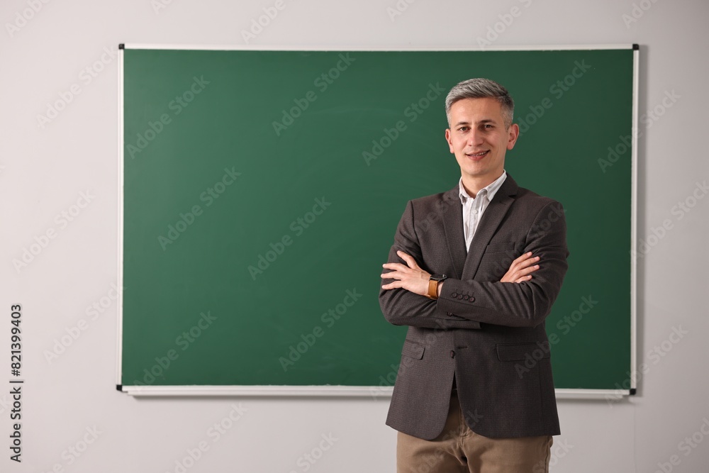 Happy teacher with crossed arms near chalkboard in classroom, space for text Happy teacher with crossed arms near chalkboard in classroom, space for text