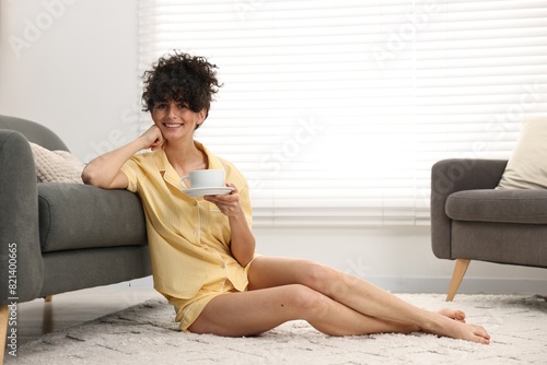 Beautiful young woman in stylish pyjama with cup of drink on floor at home