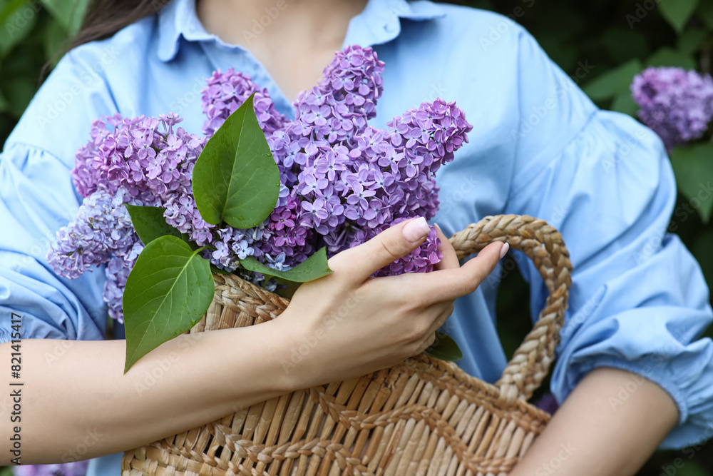Obraz premium Beautiful young woman holding wicker bag with bouquet of lilac flowers in park on spring day, closeup