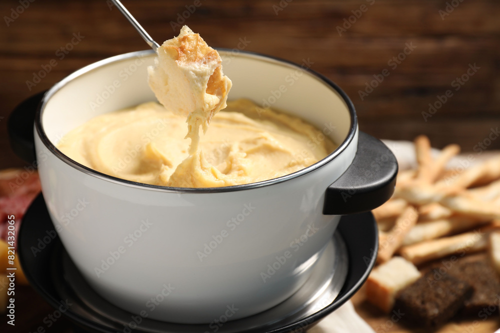 Dipping piece of bread into fondue pot with melted cheese on blurred background, closeup Dipping piece of bread into fondue pot with melted cheese on blurred background, closeup