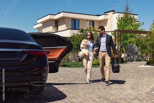 Young stylish couple hold Pomeranian Spitz in hands and walks rom their luxury suburb house to a car