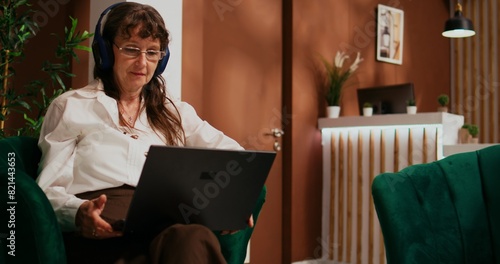 Older tourist watching movie on laptop, listening to favorite tv show in lounge area at hotel before check in. Retired woman with headset and computer to pass time on international holiday waiting. photo