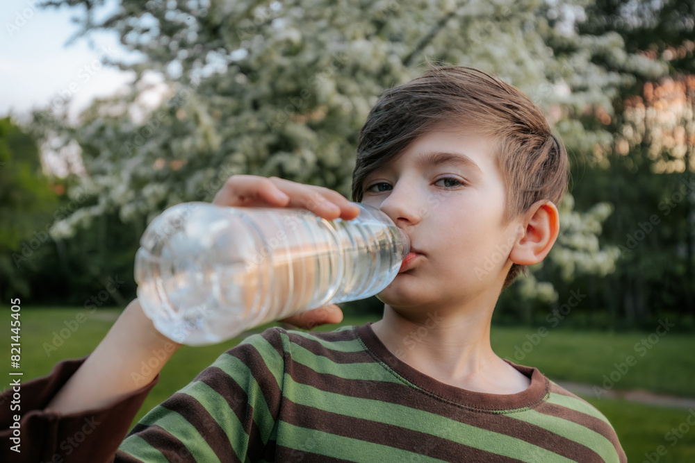 young boy drinking water from a plastic bottle, wearing a striped shirt, with a blurred green outdoor background. young boy drinking water from a plastic bottle, wearing a striped shirt, with a blurred green outdoor background.