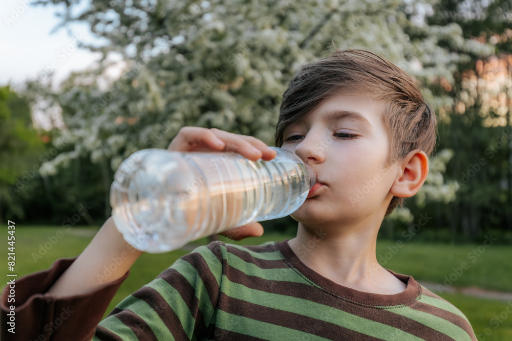 young boy drinking water from a plastic bottle, wearing a striped shirt, with a blurred green outdoor background. young boy drinking water from a plastic bottle, wearing a striped shirt, with a blurred green outdoor background.