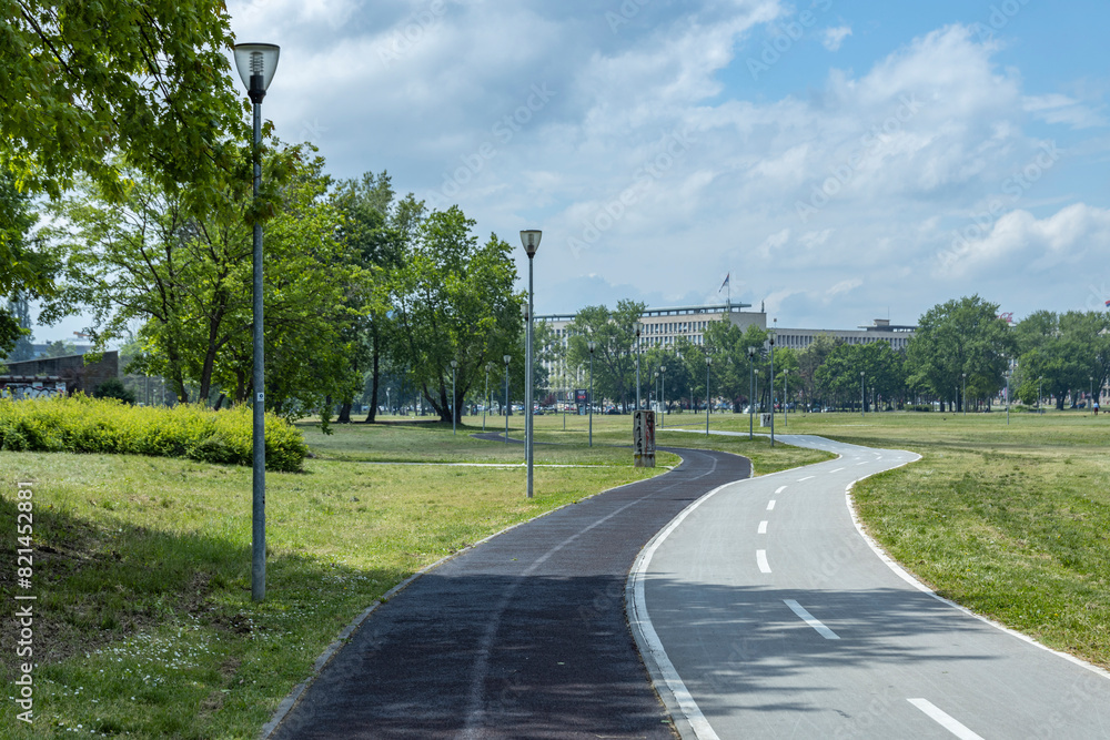 A serene park pathway with a curved asphalt road, lined with lampposts and surrounded by lush green trees and grass.