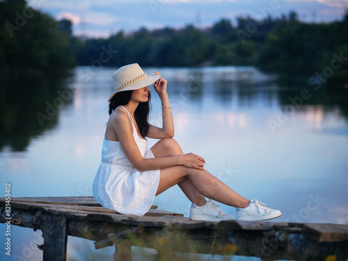 elegant fashionable young woman in a white dress and a straw hat poses by the water