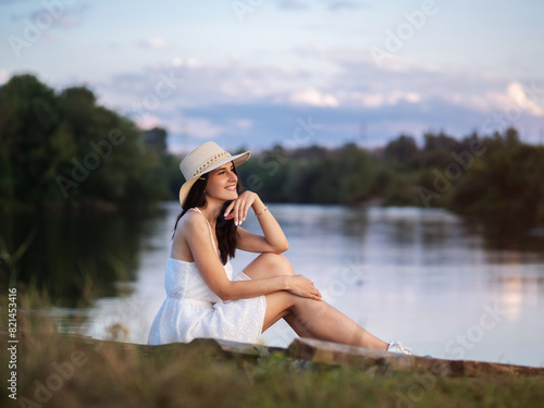 A happy smiling young woman in a white dress and a straw hat sits on the riverbank at sunset