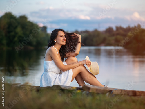 A happy smiling young woman in a white dress and a straw hat sits on the riverbank at sunset