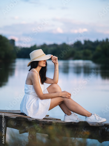 elegant fashionable young woman in a white dress and a straw hat poses by the water