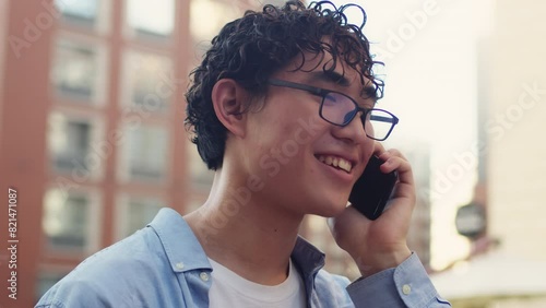 Young asian man with curly hair and glasses standing in the city street in sunny day, talking with a friend on a smartphone and cheerfully smiling close up photo