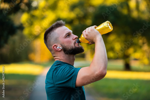 A sporty young man listening to music on wireless headphones and drinking water or nutrition juice from a bottle after a run or workout in a summer park active healthy lifestyle..