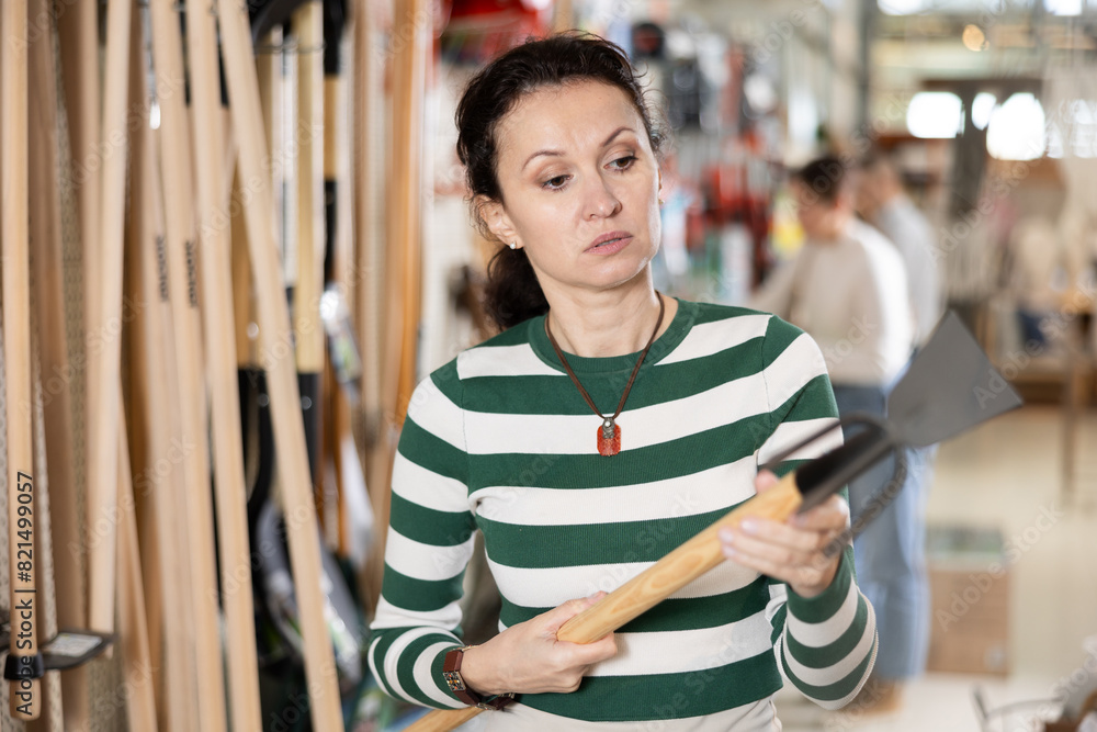 Obraz premium In garden hypermarket, woman carefully examines product before buying, chooses rake and shovel for gardening. Amateur gardener selects necessary items tools for her hobby in greenhouse flower-bed