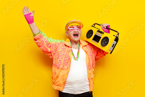 funny crazy granny in hipster clothes listening to music on tape recorder and singing on yellow isolated background, elderly cool woman dancing at party and screaming