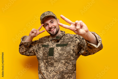 young Ukrainian army soldier in camouflage pixel uniform shows peace gesture on yellow isolated background, Ukrainian military cadet smiles and greets photo
