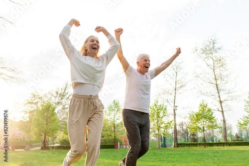 elderly couple of seniors man and woman rejoice in victory and run with raised hands in the park outdoors, gray-haired grandparents shout and celebrate luck and success and rejoice in nature
