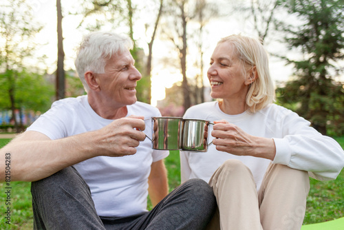 elderly couple of seniors man and woman hold metal cups and drink tea in the park outdoors, gray-haired grandparents communicate and hold mugs in nature
