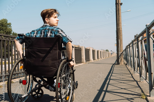 A young disabled man rides in a wheelchair across a bridge, the young male dressed in a plaid shirt and jeans.