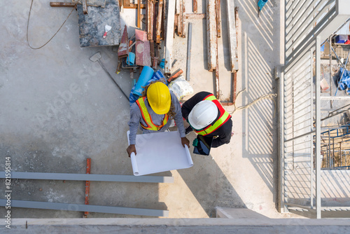 Two worker in hardhat and reflective vest are busy, looking at blueprint on a sunny day at a construction site. High angle view.