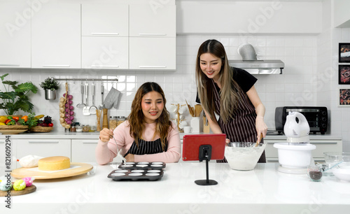 Two young women in an apron make dessert in the kitchen while looking at online recipe guide book on tablet computer.