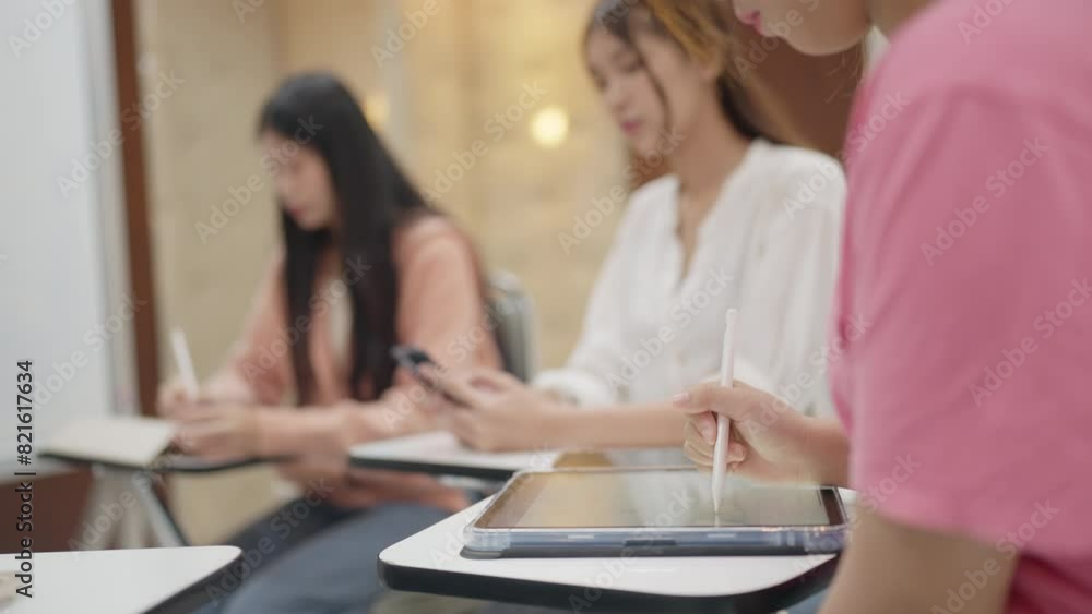 Three women are sitting at desks with a tablet in front of them. One of the women is writing with a stylus. The tablet is open to a page with a picture of a woman Three women are sitting at desks with a tablet in front of them. One of the women is writing with a stylus. The tablet is open to a page with a picture of a woman