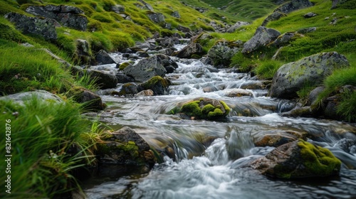 A stream of water flows through a rocky area with grass growing on the banks. The water is clear and the rocks are scattered throughout the area. The scene is peaceful and serene