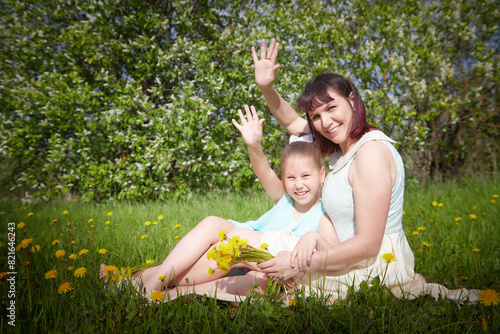 Happy mother and daughter enjoying rest, playing and fun on nature on a green lawn with dandelions and blooming apple tree on the background. Woman and girl resting outdoors in summer and spring day