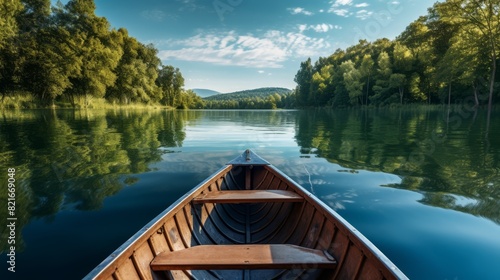 A wooden rowboat floats on a tranquil lake surrounded by lush greenery, with a clear blue sky overhead. © narak0rn