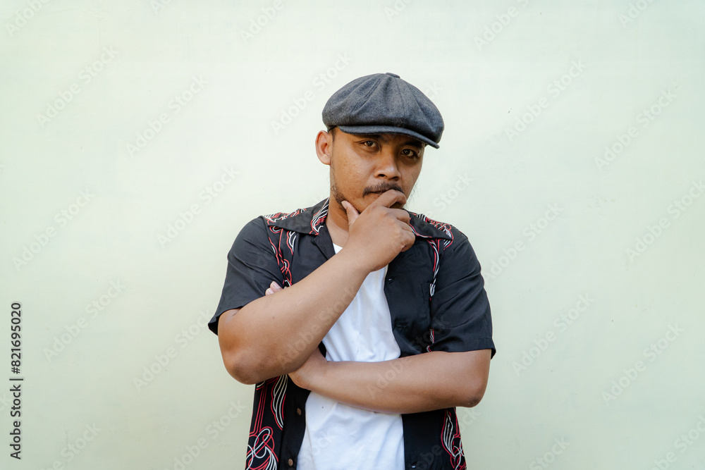 Macho man with beard and mustache looking serious isolated on beige background. Half body portrait of a vintage style Southeast Asian adult man in a patterned shirt, white tank top and newsboy cap Macho man with beard and mustache looking serious isolated on beige background. Half body portrait of a vintage style Southeast Asian adult man in a patterned shirt, white tank top and newsboy cap