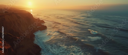 The photo shows the sunset over the ocean. The waves are crashing against the rocks. The sky is orange and the water is blue. The photo is taken from a high angle.