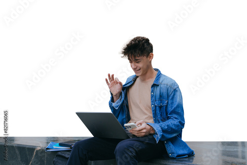 young man in a denim shirt sits outside with a laptop and phone, smiling and gesturing while engaged in a video call against transparent background