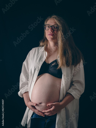 Delicate portrait of a pregnant woman on a dark background. She is wearing an open white shirt and black bra, exposing her bare belly. Her arms lovingly hugging her belly. On her legs are blue jeans.