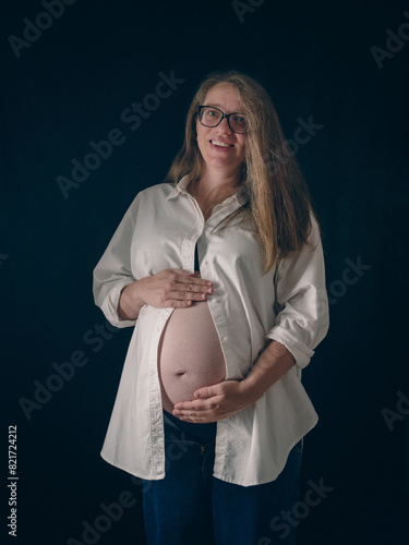 Portrait of an expectant mother lovingly cuddling her exposed belly. She is dressed in a casual white shirt and blue jeans against a dark background. Concept of joy and love of upcoming motherhood.