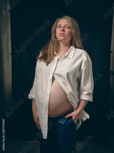 Portrait of a pregnant woman standing against a dark background. She is wearing a white unbuttoned shirt, exposing her belly, and denim trousers. Her left hand is slipped carelessly into her pocket.