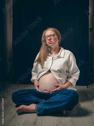 Portrait of a pregnant woman sitting cross-legged on the floor, white shirt unbuttoned, exposing her bare belly. Dramatic atmosphere of anticipation and the beauty of motherhood.